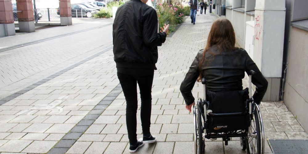 Ein junge Frau im Rollstuhl (rechts) und eine jung Frau geht daneben. Der Betrachter folgt den beiden, man sieht sie von hinten.