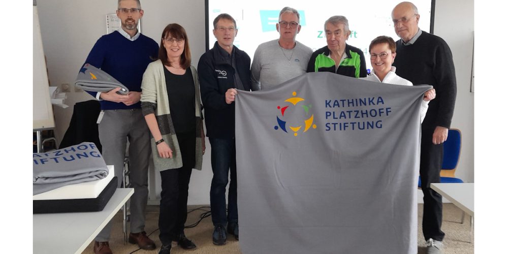Sieben Personen stehen zusammen und halten eine graue Decke in den Händen. Auf der Decke ist das Logo der Kathinka-Platzhoff-Stifung abgedruckt.
