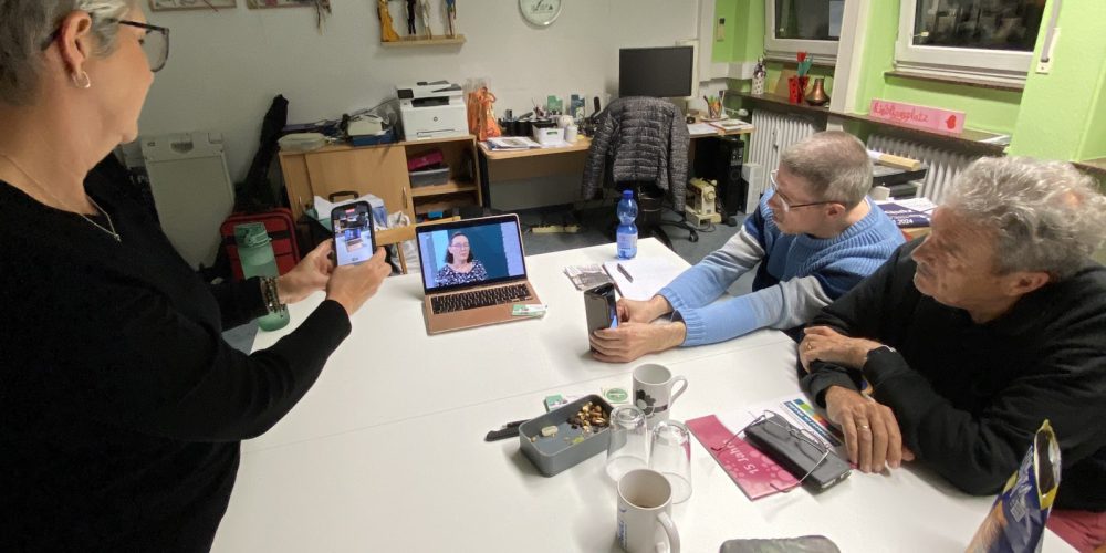 Iris, Peter und Wolfgang sind in Büro von Menschen in Hanau. Der Laptop steht auf einem Tisch. Sie schauen auf einen Laptop Sie schauen sich den Fernsehbeitrag an. Iris und Wolfgang halten Handys in den Händen und fotografieren den Bildschirm.