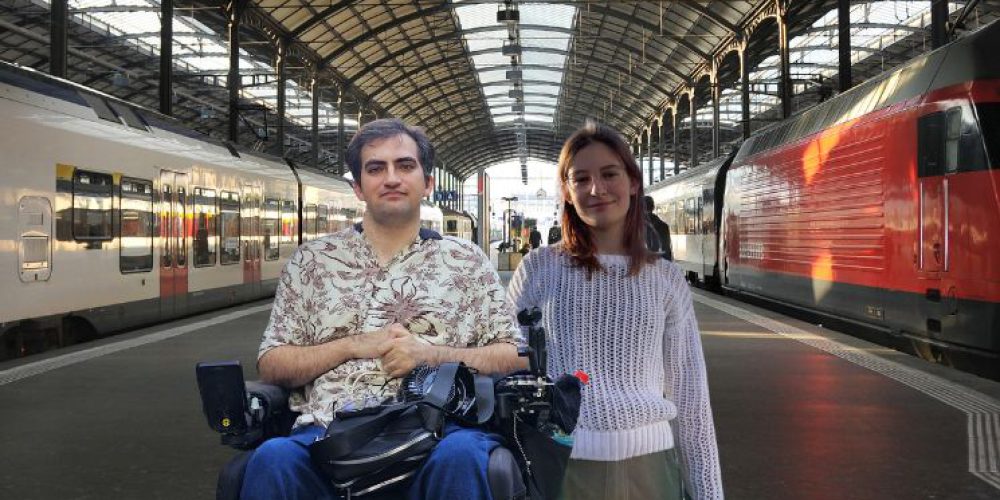 Es sind Pedram, ein junge Mann im Rollstuhl, neben Paula, einer jungen Frau mit lilanem Pullover, zu sehen, die auf einem Bahnsteig in der Mitte neben zwei Zügen stehen.
