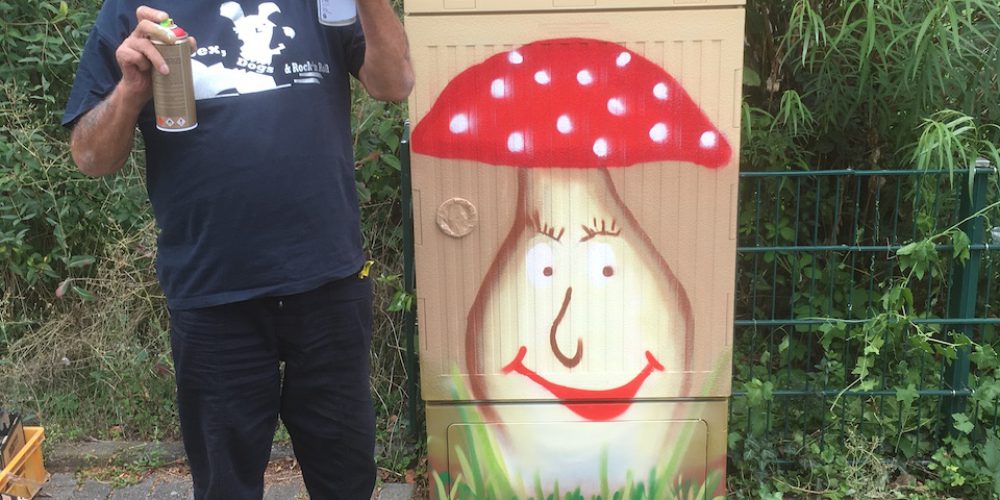 Ein Mann mit weißem Bart und Haar steht stolz mit Spraydosen neben einem Stromkasten, auf dem sich ein lächelnder Pilz befindet
