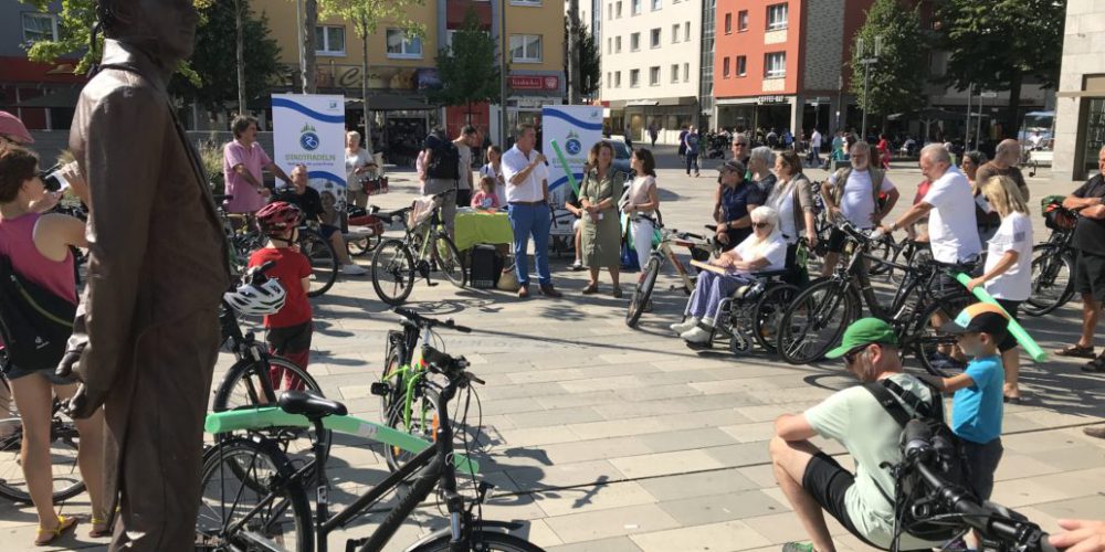 Viele Rad-Interessierte sind mit dabei. Es sind etwa 30 Männer und Frauen mit ihren Rädern zu sehen. Das Oppenheim Denkmal trägt einen Fahrradhelm.