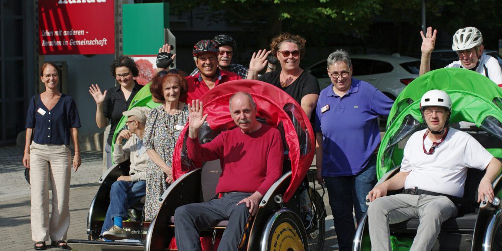 Drei Rikschas, zwei grüne und eine rote sind zu sehen. Drum herum stehen viele Menschen und heben die Hand zum Gruß.