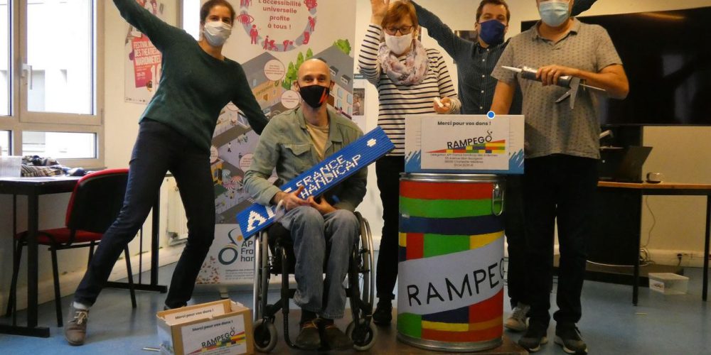 Auf dem Bild sieht man glückliche Engagierte und einen Rollstuhlfahrer in der Mitte, der eine mobile Rampe aus LEGO-Steinen in der Hand hält. Unten auf dem Boden liegen LEGO-Steine. Auf einer Tonne steht der Name des Projekts: RAMPEGO.