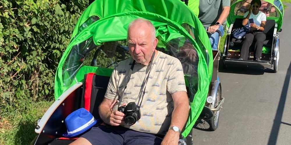 Ein Mann sitzt in einer fahrenden Rikscha mit einem grünen Dach und trägt eine Kamera um den Hals.