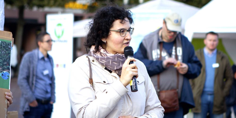 Dieses Foto von unserem Vorstansmitglied Cettina ist beim 1. Demokratie-RAUM 2019 entstanden. Sie hält in ihrer rechten Hand ein Mikrofon und kündigt einen Redner an.