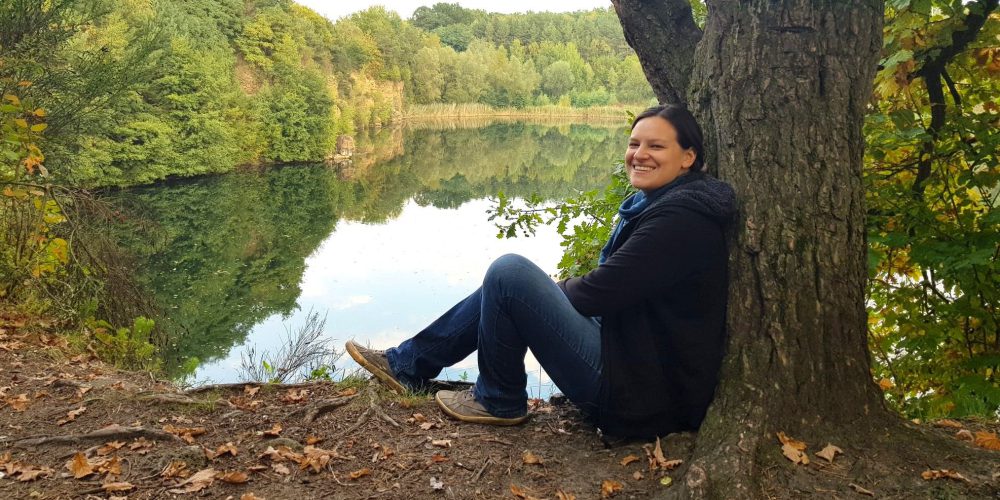 Eine junge Frau in Jeans und Sneaker sitz auf einem Baumstamm am Ufer eines Sees. Die Bäume am Seeufer spiegeln sich in der Wasseroberfläche.
