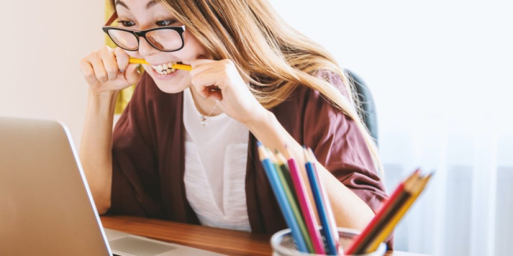 Eine Frau mit langen Haaren und Brille sitzt vor ihrem Laptop und hält mit beiden Händen einen Bleistift, in den sie hineinbeißt. Sie scheint frustriert, dass etwas nicht funktioniert.