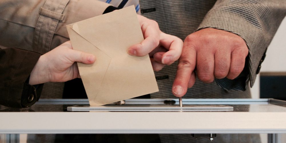 Auf dem Bild sieht man mehrere Hände, die gemeinschaftlich einen braunen Brief in einen Wahlschlitz stecken.