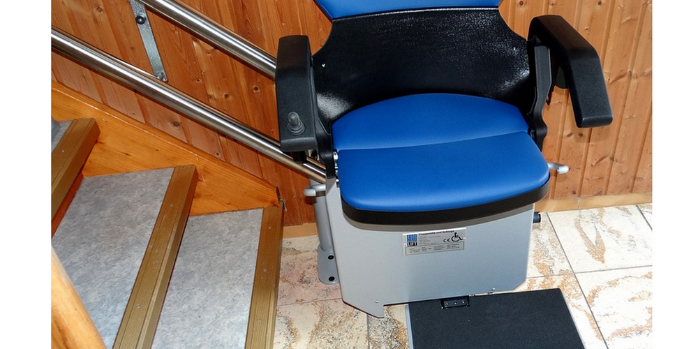 Treppenlift an einer Treppe: Ein Stuhl mit blauen Polstern und Armlehnen steht unten an einer normalen Hausflugtreppe bereit.