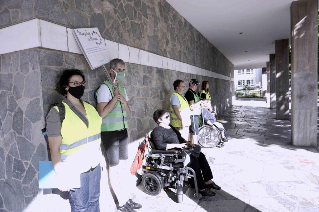 7 Personen lehnen an einer grauen Wand. Zwei von ihnen sitzen im Rollstuhl und zwei nutzen einen Langstock. Sie tragen Warnwesten. Das war eine Aktion, um Hindernisse im öffentlichen Raum sichtbar zu machen.