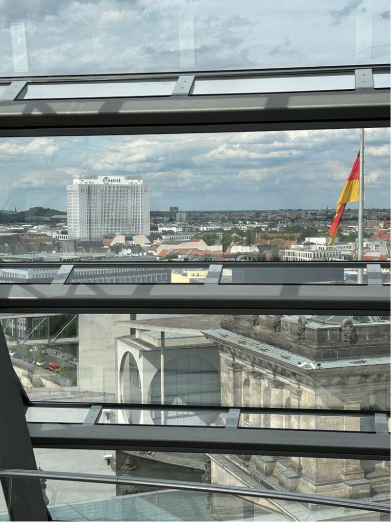 Blick durch ein Glasfenster, mit Deutschlandflagge und einem hohen Haus im Hintergrund.