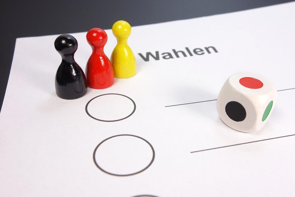Auf einem Papier mit dem Aufdruck "Wahlen" stehen 3 Holz-Spiel-Figuren in den Farben schwarz-rot-gelb auf der linken Seite. Rechts befindet sich ein Würfel mit Farbfeldern.