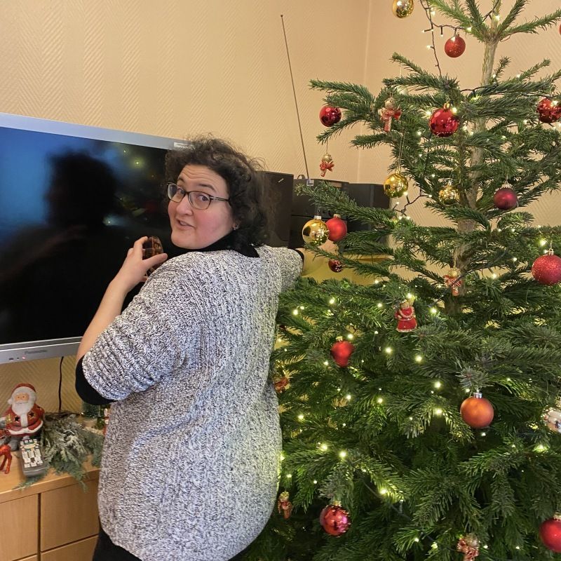 Cettina steht neben einem geschmückten Weihnachtsbaum und versucht einen CD-Spieler zum Laufen zu bekommen.