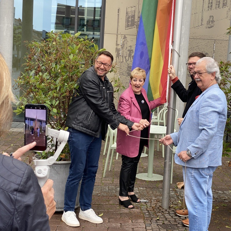 Vier Personen hissen die Regenbogenfahne zum Zeichen für Vielfalt und Zusammenhalt für alle Menschen.