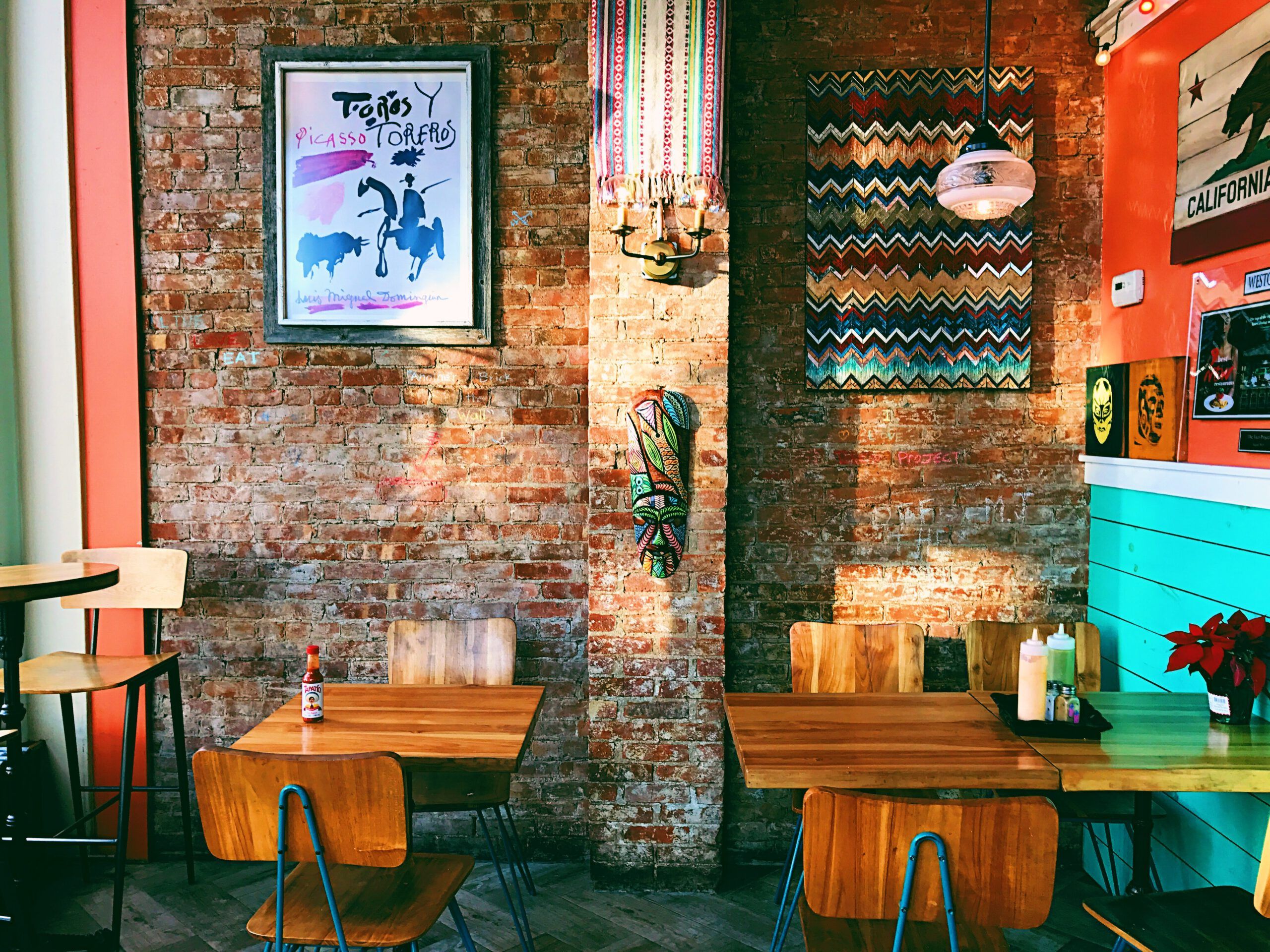 Zwei Holztische mit Süthlen vor einer Bachsteinwand in einem Café. An der Wand hängen bunte Bilder.