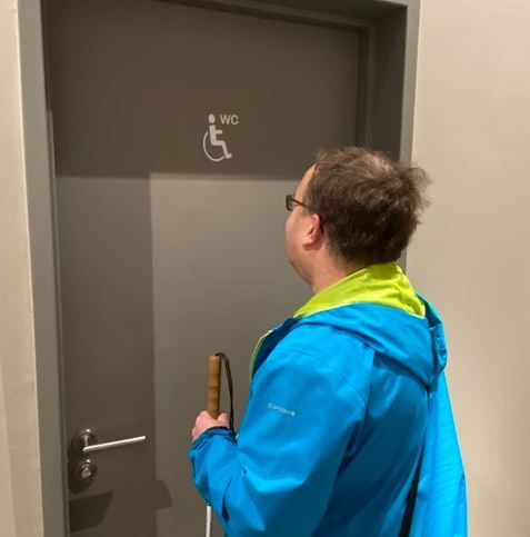 Eine Person mit einem Langstock in der Hand steht vor einer Tür, die auf Gesichtshöhe das Piktogramm eines Menschen im Rollstuhl aufgeklebt hat.