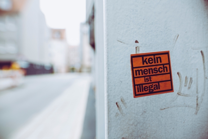 An einer weißen Wand klebt ein Aufkleber mit dem Text: Kein Mensch ist illegal