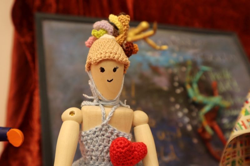 Eine Holzfigur, der ein lächelndes Gesicht aufgemalt wurde, trägt einen gehäkelten Hut, mit verschiedenen Haarfarben, die daraus hervortreten und ein blaues Kleid. Darauf ist ein großes, rotes Herz genäht.