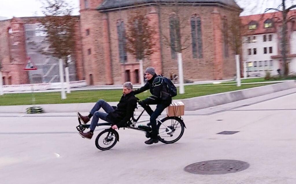 Auf dem Pino-Tandem sitzen 2 Männer und fahren vor der Wallonisch-Niederländischen Kirche