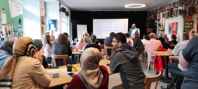 An mehreren Tischen sitzen Gruppen und diskutieren miteinander. Vor einer Leinwand steht ein Moderator und leitet die Gruppen an.