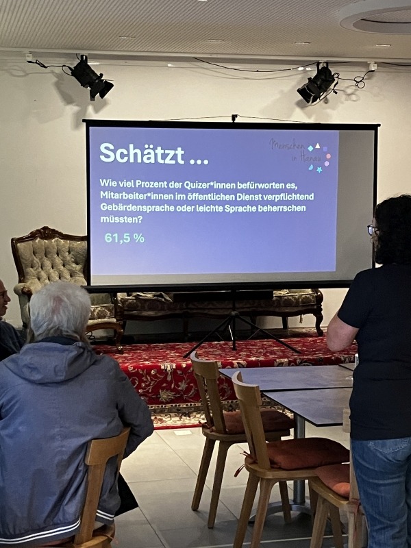 Auf einer Leinwand ist die Frage zu sehen: Schätzt...Wie viel Prozent der Quizzer sind dafür, dass im öffentlichen Dienst Gebärdensprache oder leichte Sprache von beherrscht werden müssen? Die Antwort 61,5 % der Anwesenden haben dafür gestimmt.