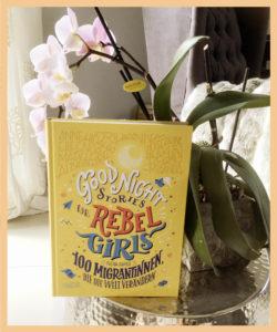 Ein Buch-Umschlag mit dem Titel Rebell Girls - 100 Migrantinnen" im Vordergrund vor einer rosafarbenen Orchidee.