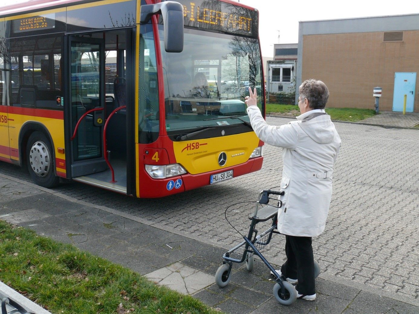 Neben einem Bus steht eine Person mit Rollator und hebt die Hand zum winken.
