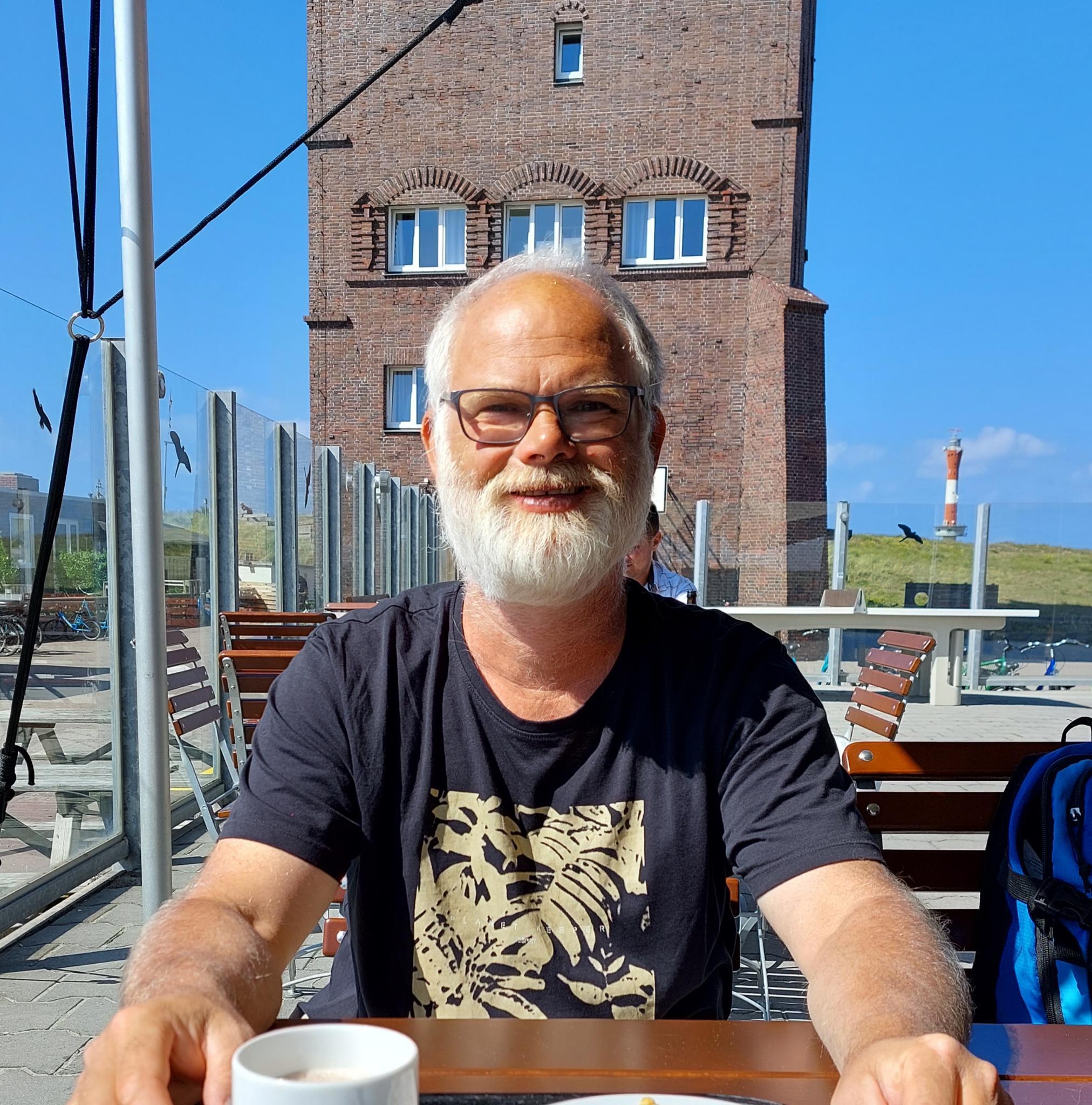 Sven mit Bart und Brille an einem sonnigen Tag vor einem Turm sitzend bei einer Tasse Kaffee