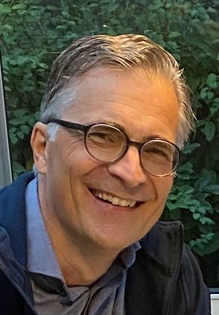 Gerhard lächelnd und mit Brille und kurzen Haaren vor einer grünen Hecke.