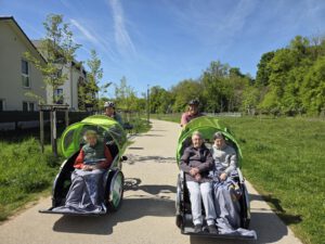 Zwei Rikschas mit Seniorinnen unter Decken fahren bei Sonnenschein auf einem Weg am Feld entlang.