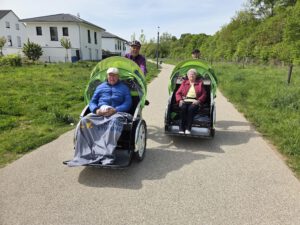 Zwei Rikschas mit Fahrgästen unter Decken fahren auf einem Weg zwischen Wiesen und Bäumen.