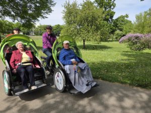 Zwei Senior:innen sitzen lächelnd in Rikschas unter einem blühenden Baum, dahinter die Pilot:innen.