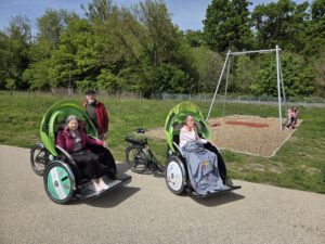 Zwei Rikschas mit Fahrgästen stehen auf einem Weg neben einem Spielplatz mit Schaukel.