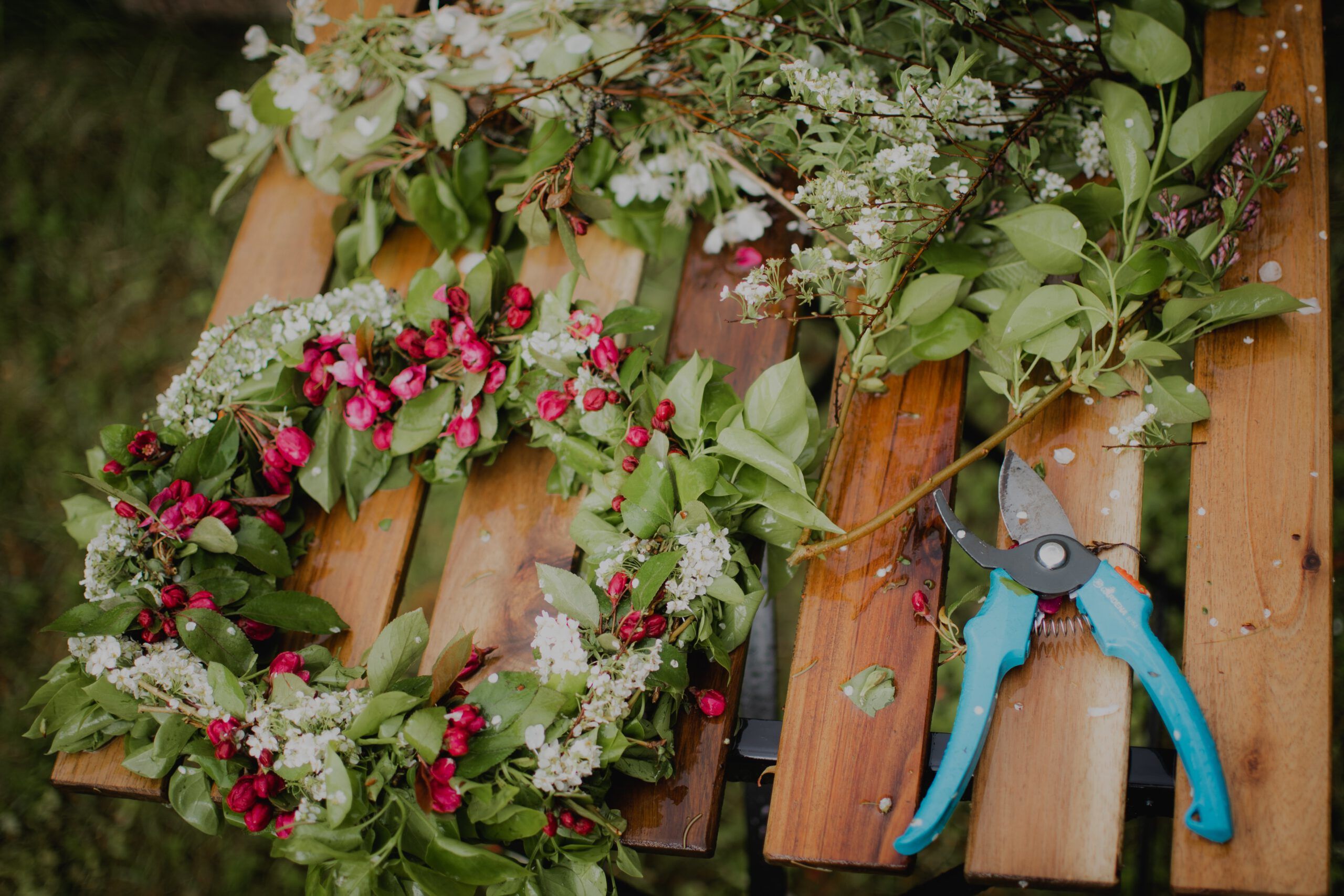 Auf dem Bild ist eine Blumenkranz auf einer Holzbank zusehen. Daneben liegt eine hellblaue Gartenschere.