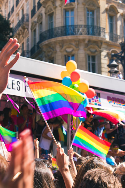Ein Bildausschnitt aus einer Pride-Parade. Verschiedene Hände schwenken Regenbogenfahnen.