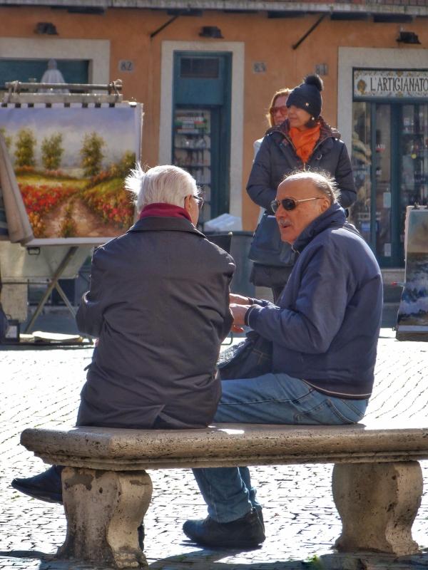 Auf einer Steinbank sitzen zwei ältere Männer und unterhalten sich lebhaft. Sie sitzen im Freien auf einem belebten Platz.