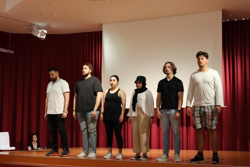 Szene aus dem interaktiven Theaterworkshop. 6 Personen stehen in einer Reihe und hören einer Erzählerin zu. Sie blicken alle nach vorne.