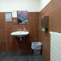 Zu sehen ist ein braun gefliester Raum von inner mit Spiegel über einem weißen Waschbecken, Papierkorb und Trockentuchspender.