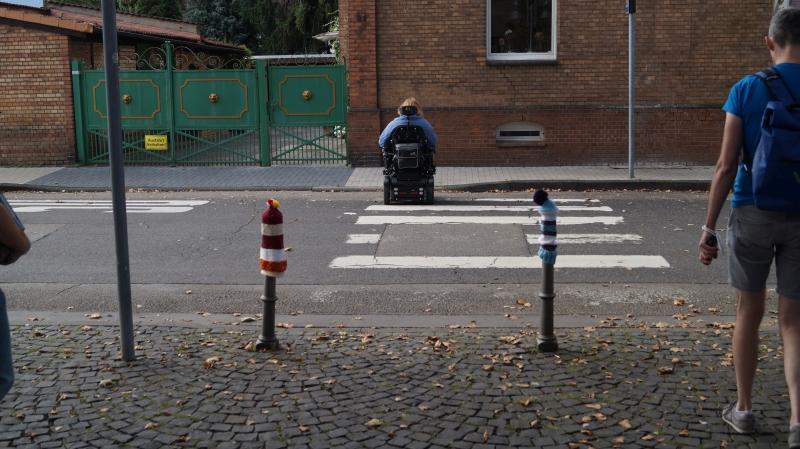 Ein schwer zu erkennender Zebrastreifen endet an einem Bürgersteig, der zur hälfte abgesenkt ist. Eine Frau mit einem E-Rollstuhl bleibt stecken. Eine unnötige Barriere.